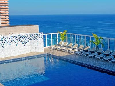 Blick auf einen rechteckigen Pool mit Liegestühlen und Palmen an der Meeresküste.