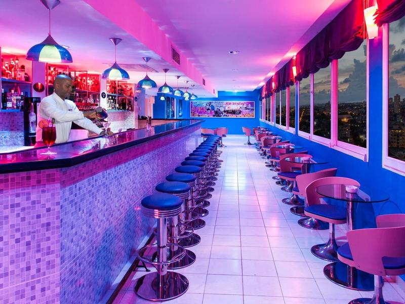 Bar d'hôtel moderne avec des tabourets bleus et des sièges roses, éclairée en bleu et rose.