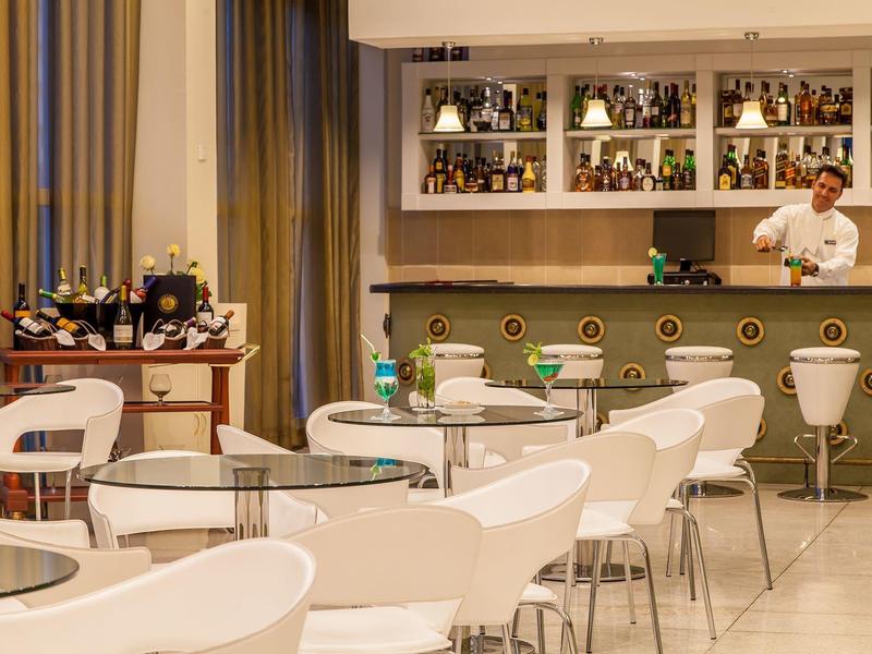 Espace bar d'hôtel moderne avec des chaises blanches, des tables et un barman derrière le bar.