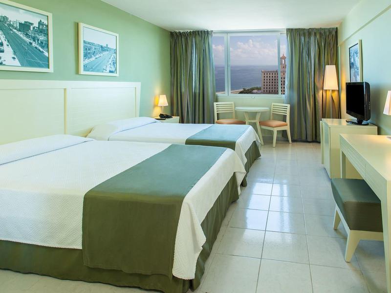 Chambre d'hôtel lumineuse avec deux lits simples, bureau, télévision et fenêtre avec vue extérieure.