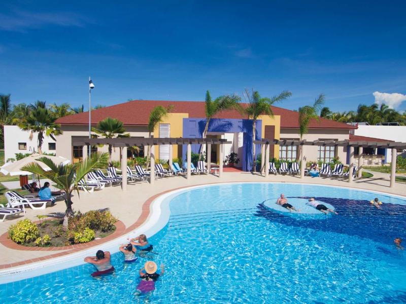 Großer blauer Pool mit runden Formen, umgeben von Liegestühlen, Palmen und Gebäuden bei Sonnenschein.