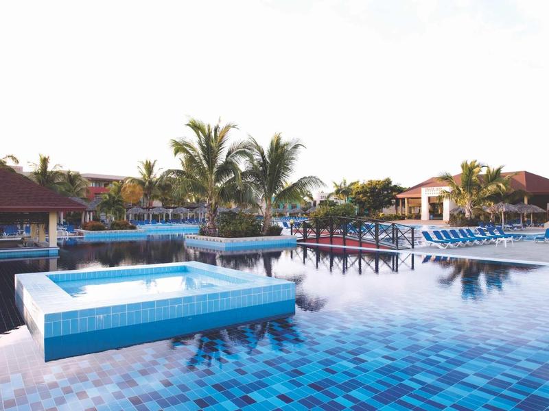 Großer Infinity-Pool mit blauen Kacheln, Sonnenliegen und Palmen in einem Resort bei klarem Himmel.