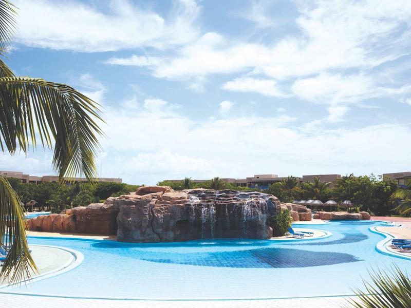 Großer Pool mit Wasserfall, umgeben von Palmen und Sonnenliegen bei blauem Himmel.