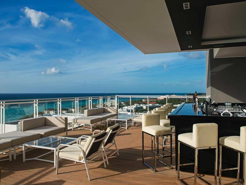 Terrazza moderna dell'hotel con bar, posti a sedere e vista sull'oceano sotto un cielo azzurro.