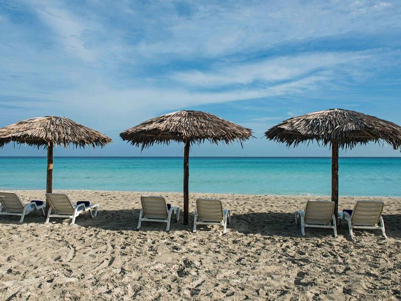 Spiaggia con tre ombrelloni di paglia e lettini davanti al mare calmo.