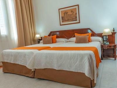 Světle zařízený dvoulůžkový pokoj s velkou postelí, oranžovými akcenty a dvěma nočními stolky.