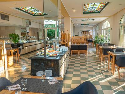 Světelná hotelová restaurace s vzorovanou podlahou, dřevěnými židlemi a velkými okny.