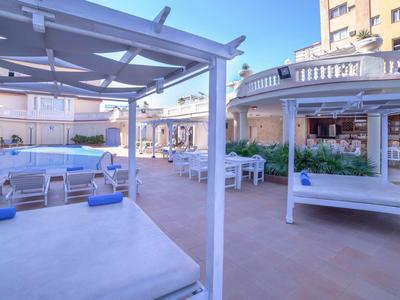 Světlá terasa s denními postelemi a židlemi, obklopená budovami a sluneční clonou.