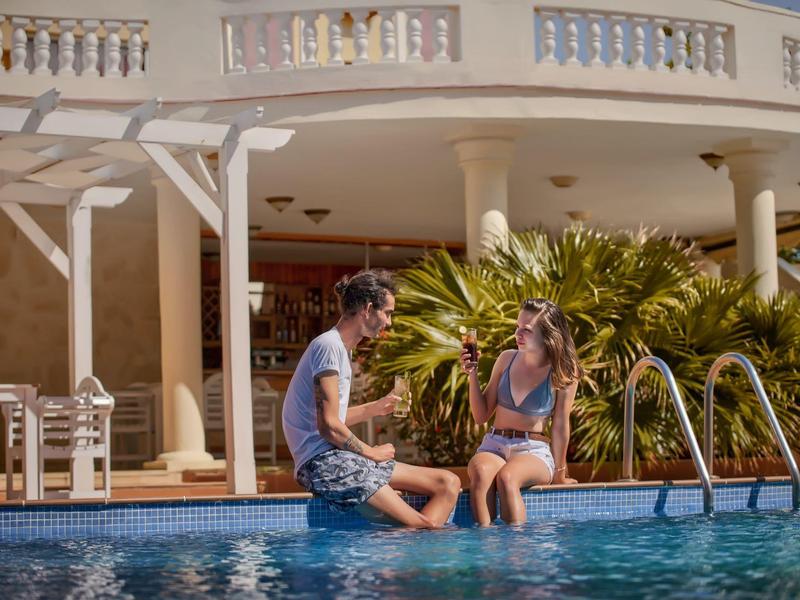 Pár sedí u okraje hotelového bazénu s palmami v pozadí.