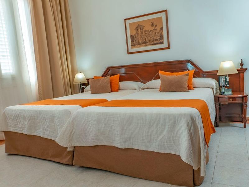 Světle zařízený dvoulůžkový pokoj s velkou postelí, oranžovými akcenty a dvěma nočními stolky.