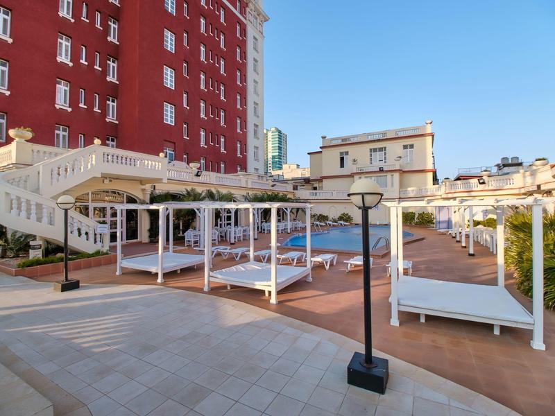 Venkovní prostor s bílými lehátky pod pergolami vedle červené budovy za jasné oblohy.