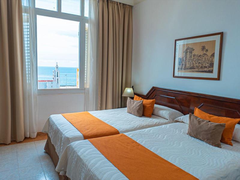 Světly dvojlůžkový pokoj s oranžovými doplňky a výhledem na moře přes velké okno.