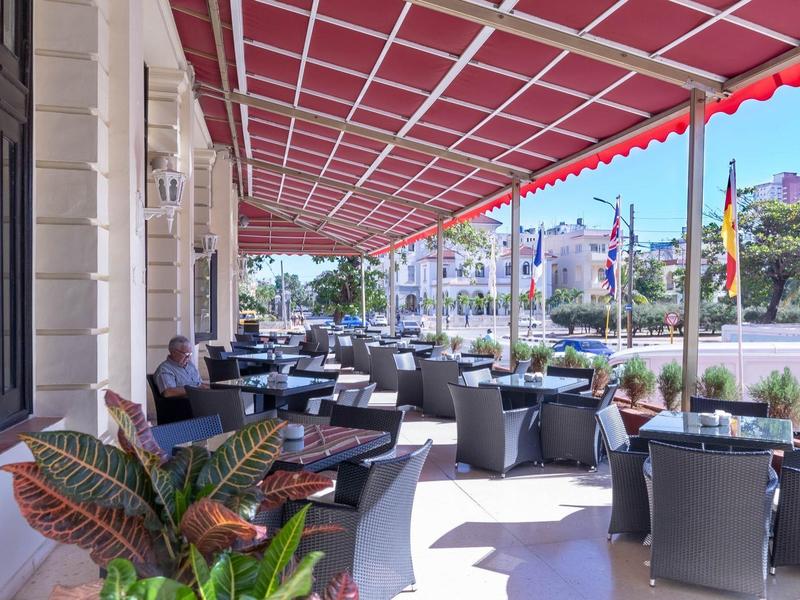 Slunečná kavárenská terasa s stoly, židlemi a červenými markýzami podél ulice.