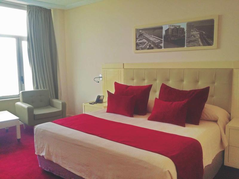 Chambre avec grand lit, coussins rouges, chemin de lit, chaise et fenêtre