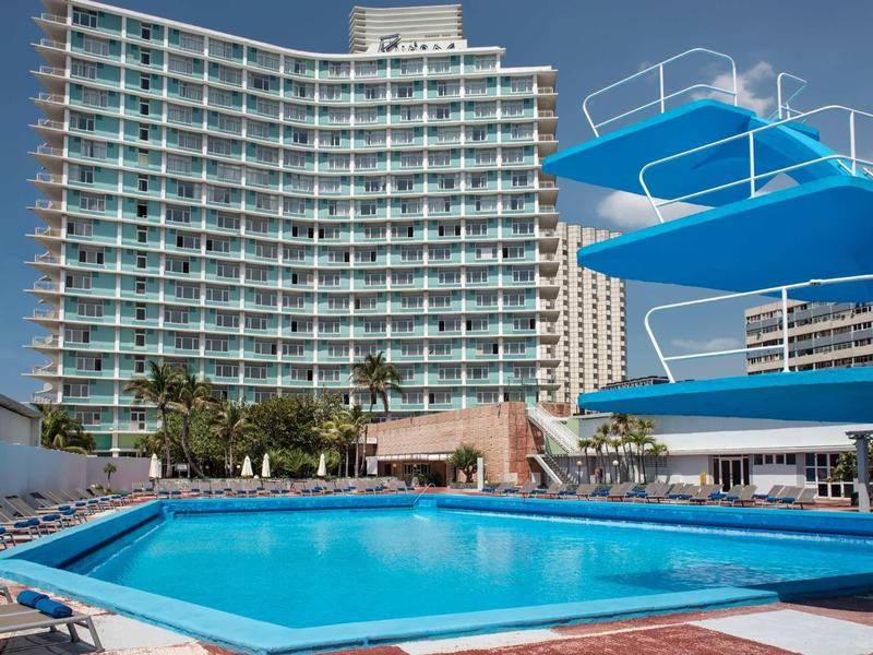 Complexe hôtelier moderne avec grande piscine extérieure et plongeoirs bleus sous un ciel dégagé.