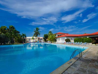 Großer Außenpool mit klar blauem Wasser, umgeben von Palmen und weißen Gebäuden unter blauem Himmel.