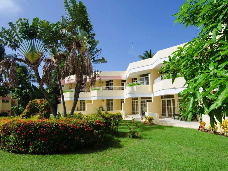 Hotel con giardino curato, aiuole di fiori e palme sotto un cielo azzurro limpido.