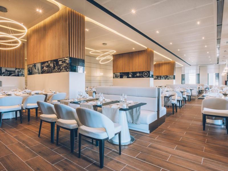 Área de restaurante de hotel moderno con suelo de madera, mesas blancas y sillas elegantes bajo luces circulares de techo.