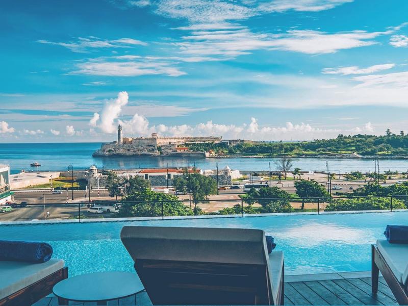 Vista de una piscina con tumbonas y un puerto al fondo bajo un cielo azul.