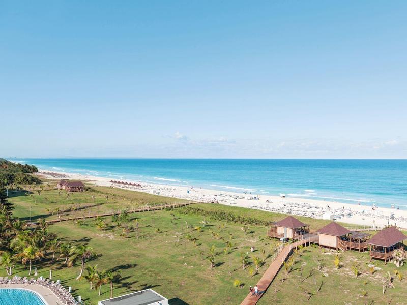 Spiaggia con sabbia chiara, mare calmo e due gazebo in legno sull'erba verde.