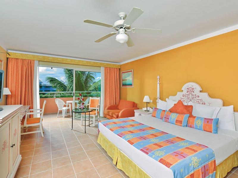 Camera d'hotel luminosa con letto colorato, divano, TV e balcone con vista su palme.