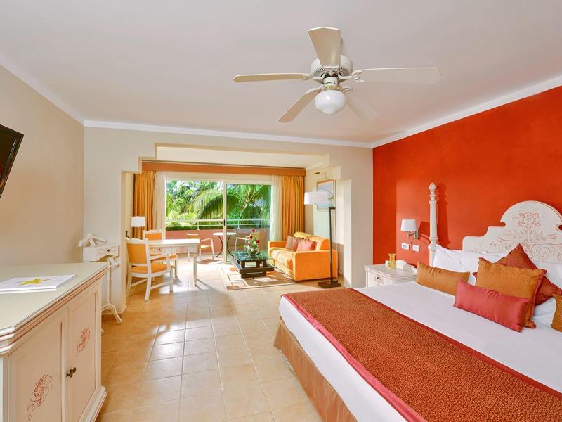 Camera d'hotel luminosa con letto grande, parete rossa, ventilatore a soffitto e balcone con vista su palme.