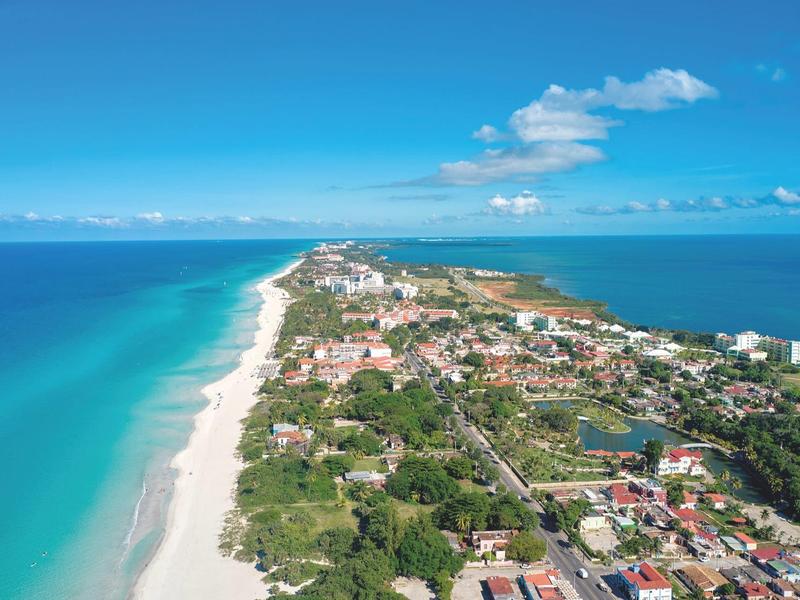 Vista aerea di una città lungo una spiaggia di sabbia bianca con mare turchese e cielo limpido.