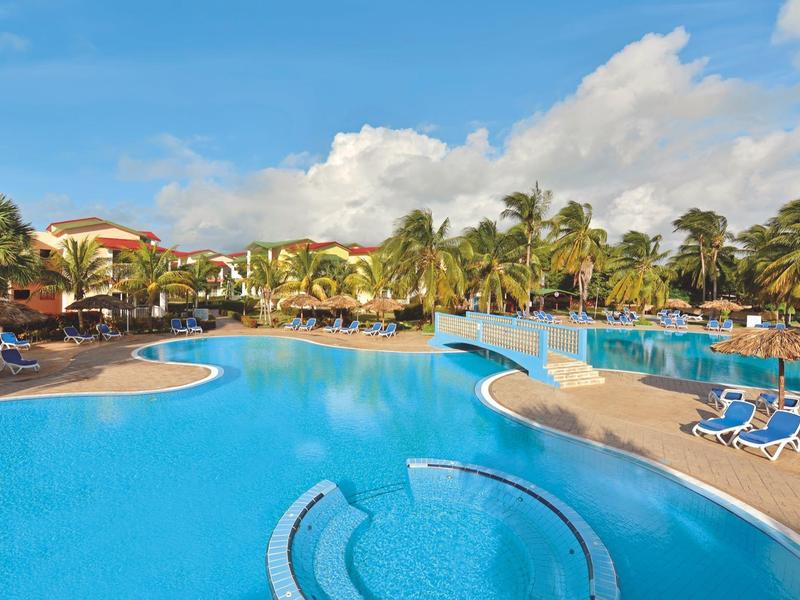 Grande piscina dell'hotel con palme e sedie a sdraio blu sotto un cielo azzurro.