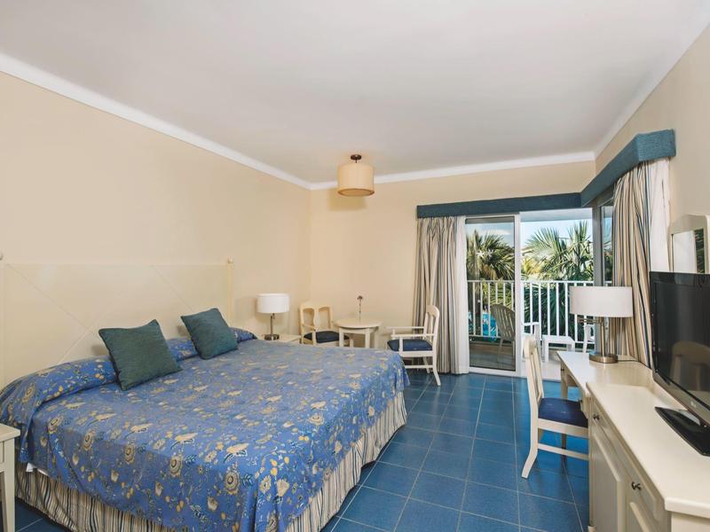 Camera d'hotel luminosa con letto matrimoniale, biancheria da letto blu e balcone con vista su palme.