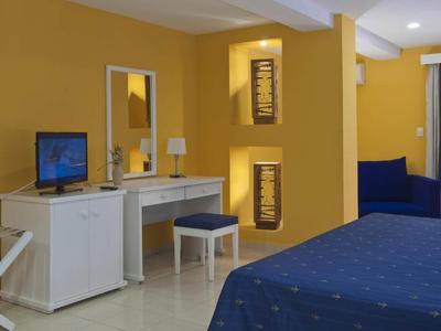 Piccola camera d'hotel con pareti gialle, letto blu e scrivania con specchio.