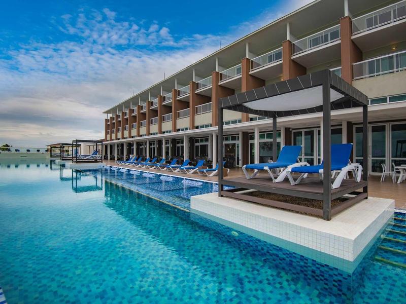 Complesso alberghiero moderno con piscina a sfioro e terrazza soleggiata sotto un cielo blu.