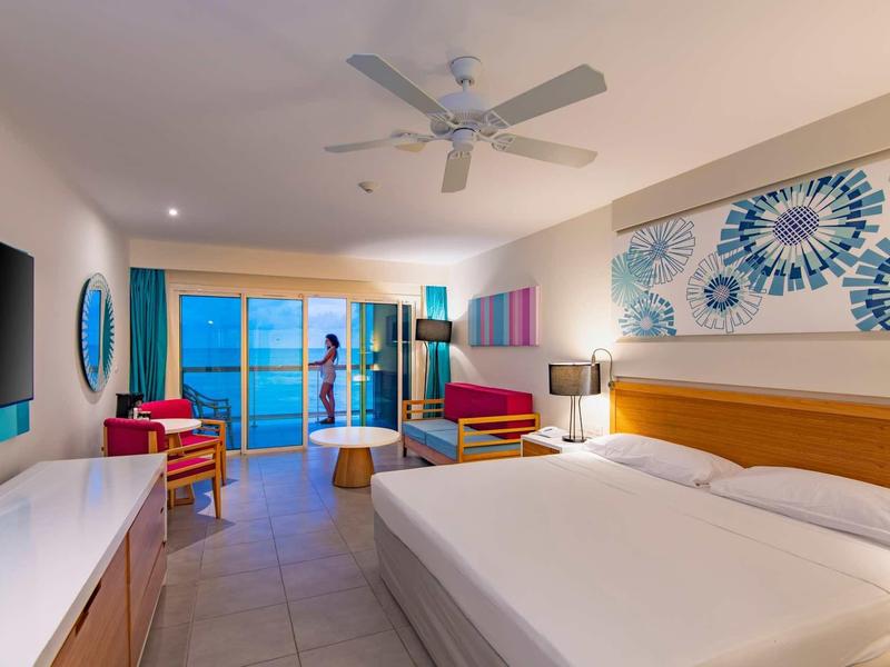 Camera d'hotel luminosa con letto matrimoniale, zona soggiorno e balcone con vista mare.