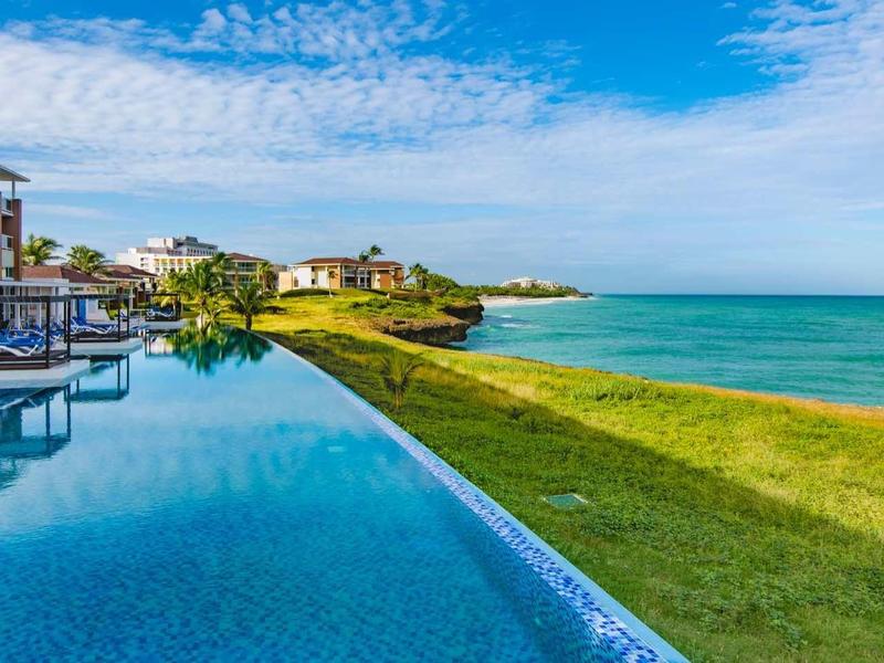 Piscina a sfioro accanto a un hotel moderno con vista su prato verde e mare blu sotto un cielo azzurro.