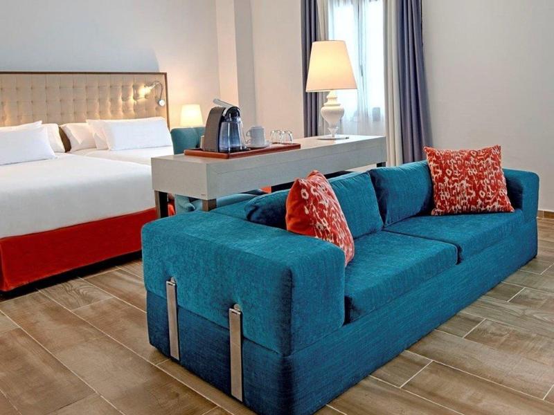 Moderne hotelkamer met een blauwe bank, bureau en groot bed met rode rand.
