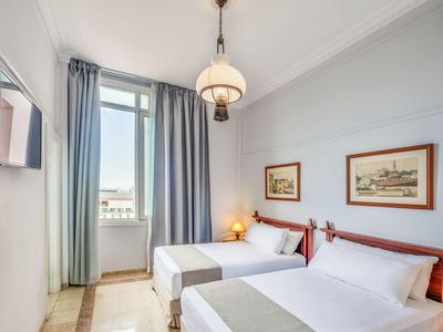 Chambre d'hôtel lumineuse avec deux lits, accès au balcon et tableaux encadrés au mur.