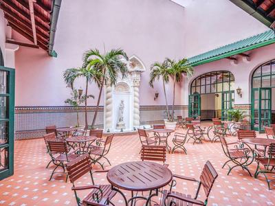 Cour intérieure d'hôtel avec terrasse, tables, chaises et palmiers sous un ciel rose pâle.