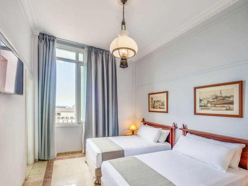 Chambre d'hôtel lumineuse avec deux lits, accès au balcon et tableaux encadrés au mur.