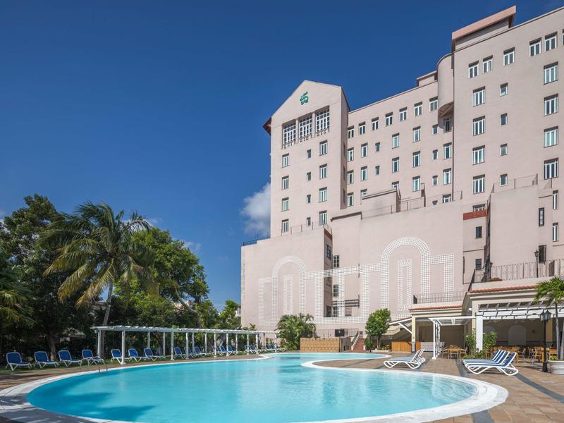 Bâtiment d'hôtel avec grande piscine extérieure, palmiers et chaises longues sous un ciel clair