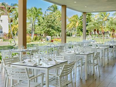 Area pranzo aperta con tavoli e sedie bianchi, circondata da palme e vegetazione.