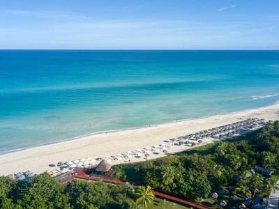 Spiaggia con sabbia bianca, lettini e acqua turchese chiara accanto a vegetazione verde.