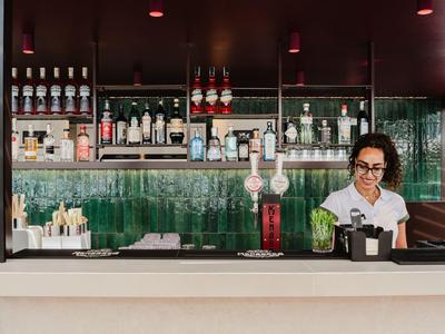 Eine Bar mit Regalen voller Flaschen und einer Barkeeperin, die Getränke vorbereitet.