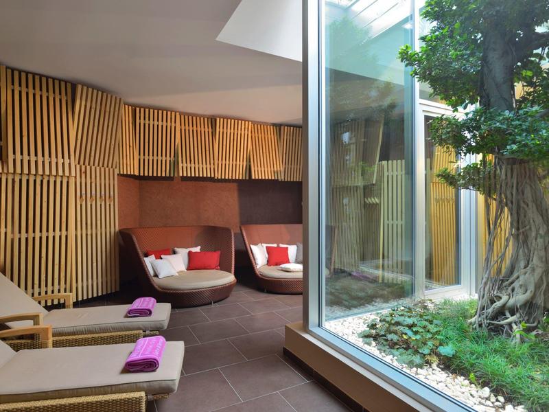 Ein moderner Ruheraum mit Liegen, Sesseln und Fenster mit Blick auf einen Garten mit Baum.