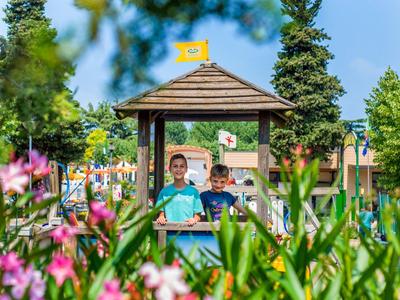 Two children sit on a swing in a playhouse in the park, surrounded by flowers.