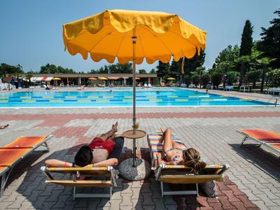 Two people relax under a large yellow umbrella by a pool with orange loungers.