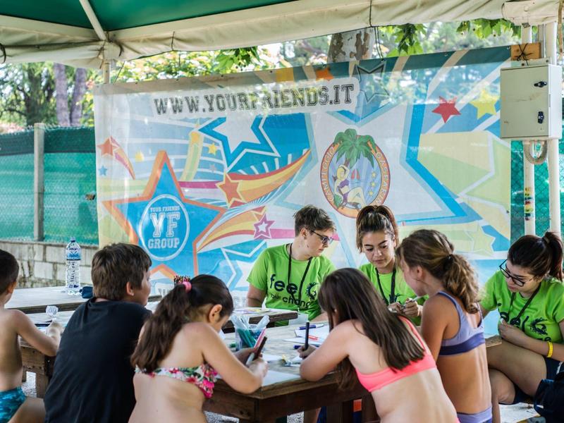 Children sit at a table under a pavilion with a colorful banner featuring logos in the background.