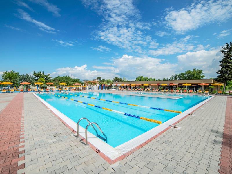 Large outdoor pool with lanes, surrounded by loungers and umbrellas under a blue sky.