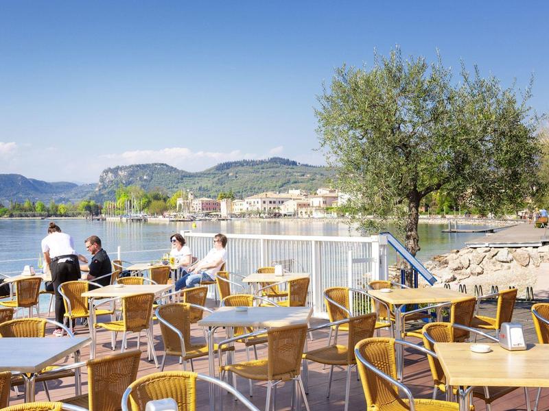 Terrazza con tavoli e sedie vicino a un lago con vista sulle montagne e ulivi.