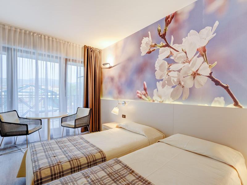 Camera d'hotel luminosa con due letti singoli, decorazione floreale a parete e due sedie vicino alla finestra.