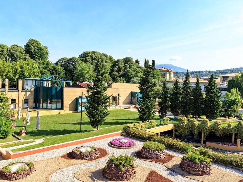 Giardino verde dell'hotel con vari cespugli, fiori, sentieri e cielo azzurro.