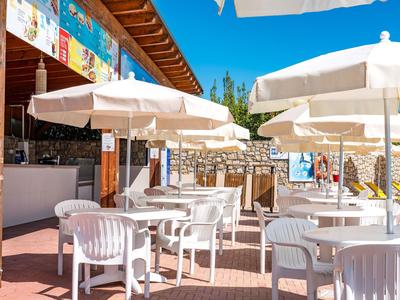 Area all'aperto con sedie bianche e ombrelloni accanto a un bar sotto un cielo azzurro.